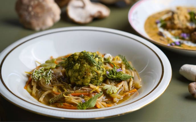Verschiedenes Julienne-Gemüse mit Saft in einem tiefen Teller mit Goldrand angerichet. Mit Kräutern garniert und in der Mitte ist ein Kräuter-Semmelknödel auf das Gemüse gesetzt.