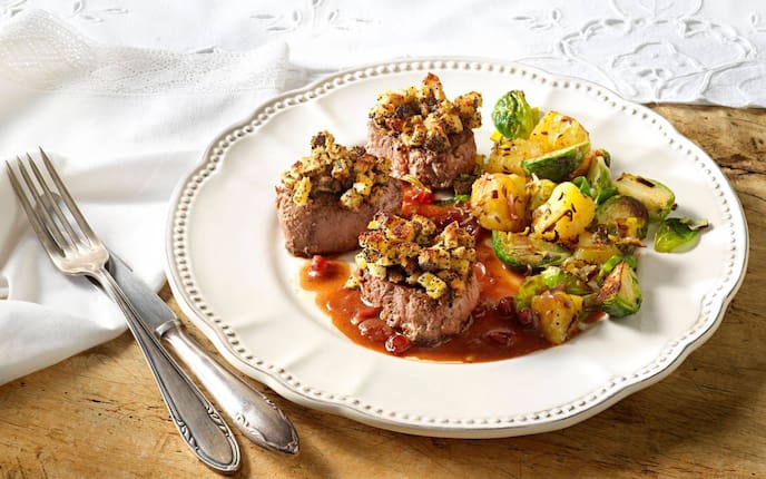 Weißer Keramikspeiseteller auf dem gebratene Hirschmedaillons mit einer Semmelwürfel-Mohn-Kruste und Erdäpfel-Rosenkohl-Beilage und Saft. Daneben liegt Messer und Gabel, alles steht auf einem alten Holztisch mit einer weißen Tischdecke samt Stoffserviette.