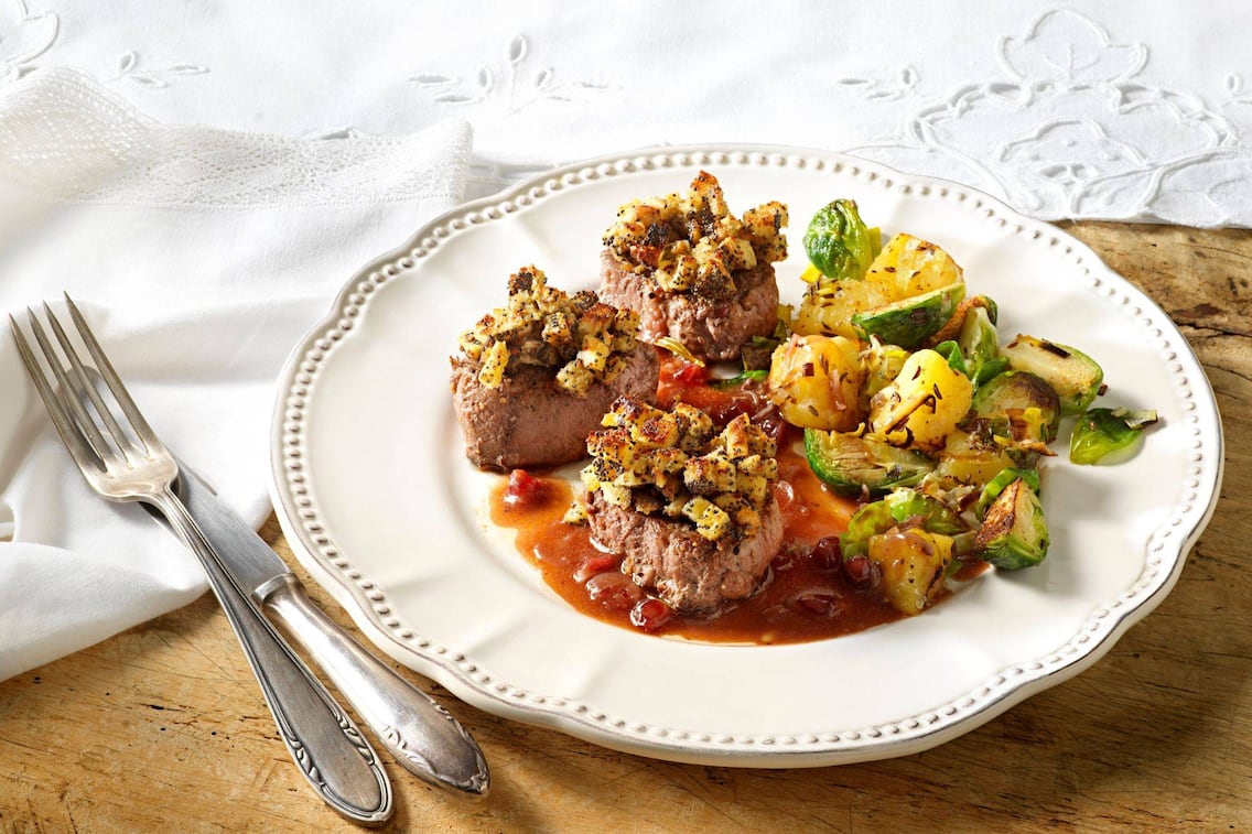 Weißer Keramikspeiseteller auf dem gebratene Hirschmedaillons mit einer Semmelwürfel-Mohn-Kruste und Erdäpfel-Rosenkohl-Beilage und Saft. Daneben liegt Messer und Gabel, alles steht auf einem alten Holztisch mit einer weißen Tischdecke samt Stoffserviette.