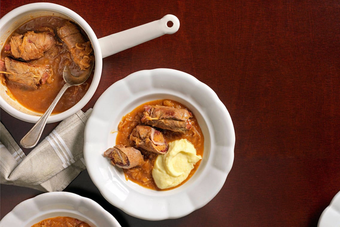 Rouladen mit Sauerkraut gefüllt, in einer roten Sauce und Erdäpfelpüree, in einer weißen Emailkaserolle mit einem alten Esslöffel und auch auf einem weißen schweren, geschwungenen Keramikteller angerichtet.