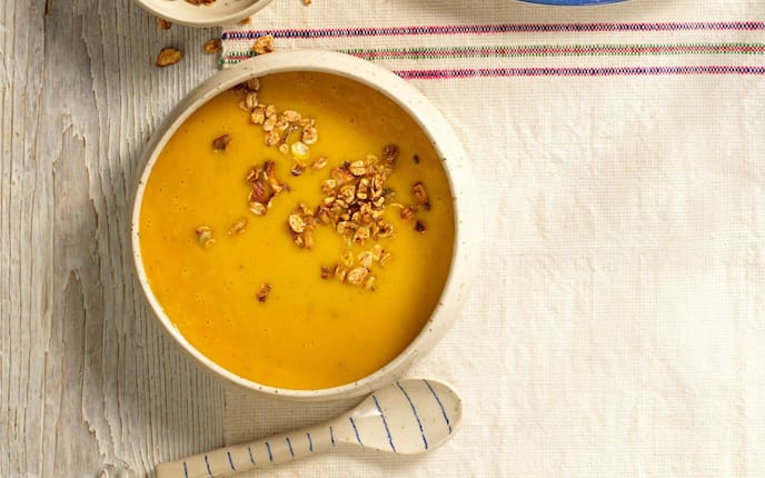 Cremige Kürbissuppe mit knusprigem Müsli in einer Suppeschüssel mit Keramiklöffel und Tischuntersetzer