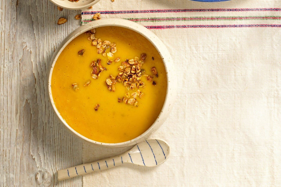 Cremige Kürbissuppe mit knusprigem Müsli in einer Suppeschüssel mit Keramiklöffel und Tischuntersetzer