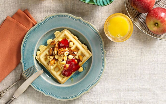 Rahmwaffeln, mit Pekannüssen, Haselnüssen, Walnüssen und Mandeln und Marmelade. Auf einem blau glasiertem Dessertteller angerichtet mit Besteck, einer orangen Stoffserviette und einem Glas Orangensaft