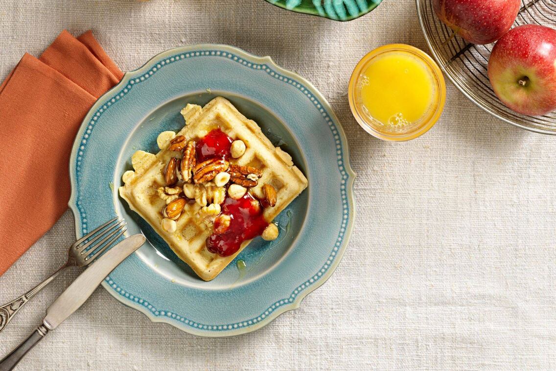 Rahmwaffeln, mit Pekannüssen, Haselnüssen, Walnüssen und Mandeln und Marmelade. Auf einem blau glasiertem Dessertteller angerichtet mit Besteck, einer orangen Stoffserviette und einem Glas Orangensaft