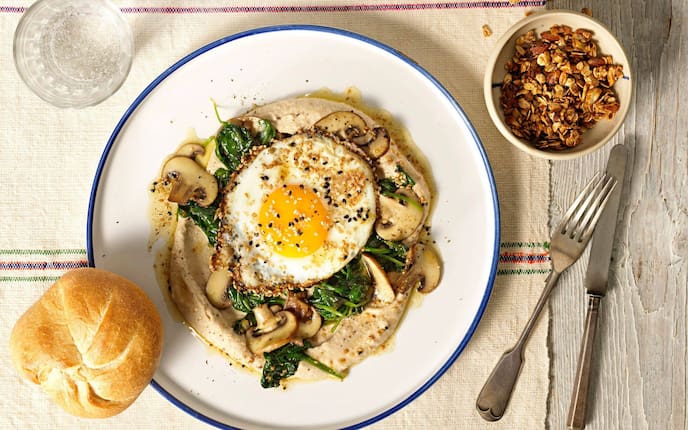 Bohnencreme mit gedünsteten Spinatblättern, geschnittenen gerösteten Champignons darauf ein Spiegelei mit hellem und dunkel Sesam. Auf einem weißen Teller mit blauem Ran angerichtet. Danaben liegt eine Kaisersemmel, Besteck, ein Glas Wasser und eine kleine Keramikschale mit Knuspermüsli