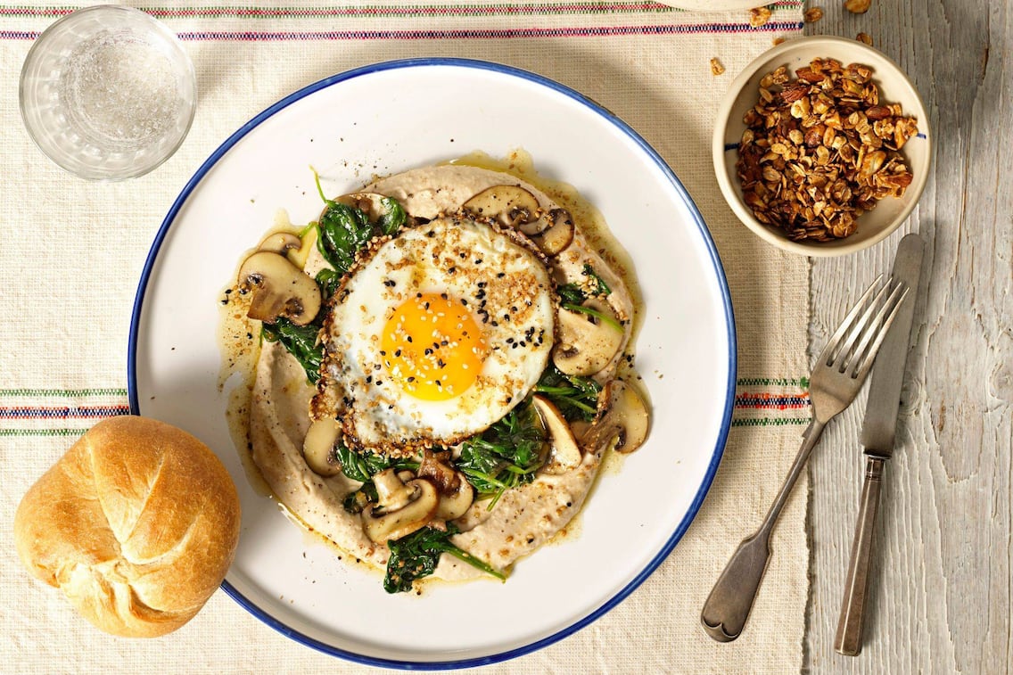 Bohnencreme mit gedünsteten Spinatblättern, geschnittenen gerösteten Champignons darauf ein Spiegelei mit hellem und dunkel Sesam. Auf einem weißen Teller mit blauem Ran angerichtet. Danaben liegt eine Kaisersemmel, Besteck, ein Glas Wasser und eine kleine Keramikschale mit Knuspermüsli