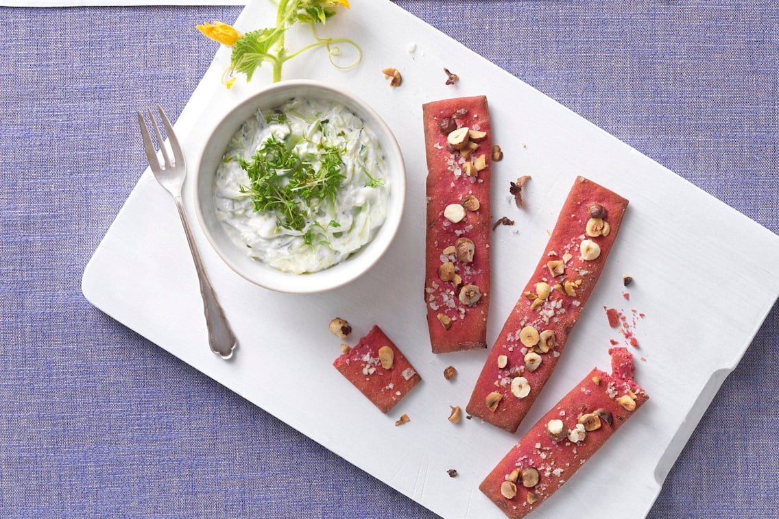 in einem weißen Schüsselchen ist der Gurkenaufstrich mit Brunnenkresse bestreut angerichtet. Das Schüsserl steht auf einem weißen Holzbrett, darauf liegt eine Dessertgabel und rotes Knäckebrot, das mit Salz und Haselnüssen bestreut ist. Alles zusammen steht auf einem blauen Tischtuch und ist von oben fotografiert.