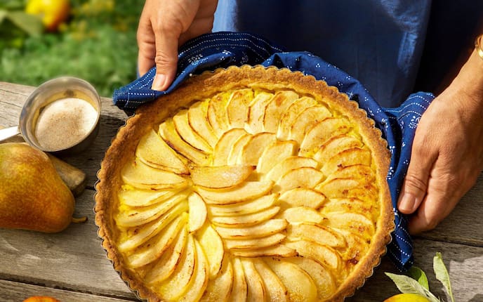 Holztisch von oben fotografiert, darauf hält ein Mann (nur die Hände sind zu sehen) einen gebackenen Kuchen auf dem in Reih und Glied Apfel- und Birnenspalten geschlichtet sind in Richtung Kamera. Danebben liegen frische Birnen und Äpfel. Der Mann hält den Kuchen mit einem Blaudrucktuch.
