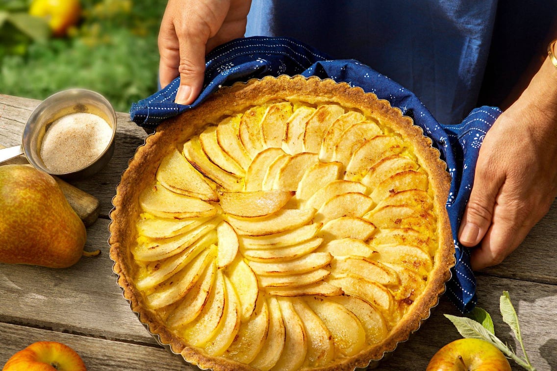 Holztisch von oben fotografiert, darauf hält ein Mann (nur die Hände sind zu sehen) einen gebackenen Kuchen auf dem in Reih und Glied Apfel- und Birnenspalten geschlichtet sind in Richtung Kamera. Danebben liegen frische Birnen und Äpfel. Der Mann hält den Kuchen mit einem Blaudrucktuch.
