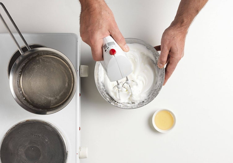 Eischnee wird mit einem Mixer aufgeschlagen, daneben steht ein Topf mit Sirup auf einer Herdplatte