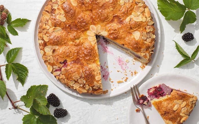 Kuchen gefüllt mit Brombeeren und mit Mandelsplitter gebacken auf einem Teller, ein Stück ist herausgeschnitten und liegt auf einem separaten Teller mit einer Dessertgabel und ein Stück ist abgestochen.