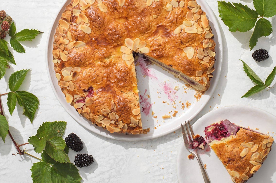Kuchen gefüllt mit Brombeeren und mit Mandelsplitter gebacken auf einem Teller, ein Stück ist herausgeschnitten und liegt auf einem separaten Teller mit einer Dessertgabel und ein Stück ist abgestochen.