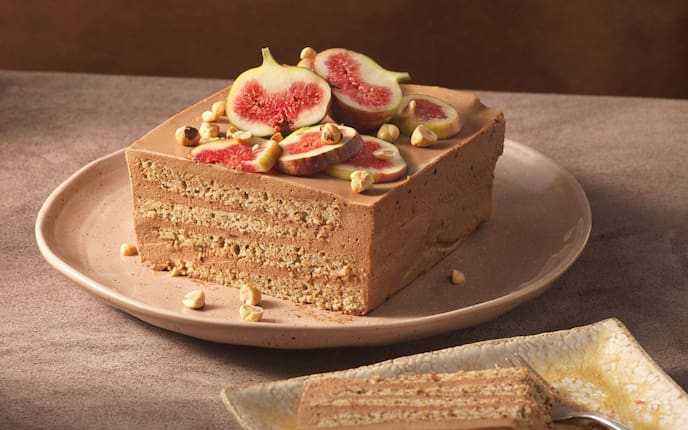 Eine Stück Torte mit dünnen Teigschichten und einer Cremefüllung, obenauf liegen ganze Haselnüsse und Feigen in Scheiben geschnitten.