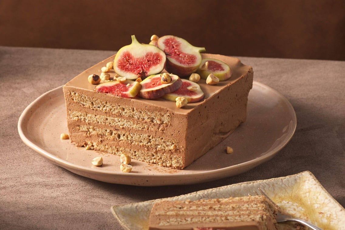 Eine Stück Torte mit dünnen Teigschichten und einer Cremefüllung, obenauf liegen ganze Haselnüsse und Feigen in Scheiben geschnitten.