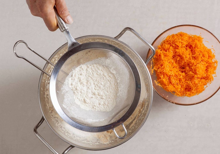Mehl wird durch ein Sieb in die Schüssel zum Teig gesiebt, daneben steht eine Glasschüssel mit geriebenen Karotten.