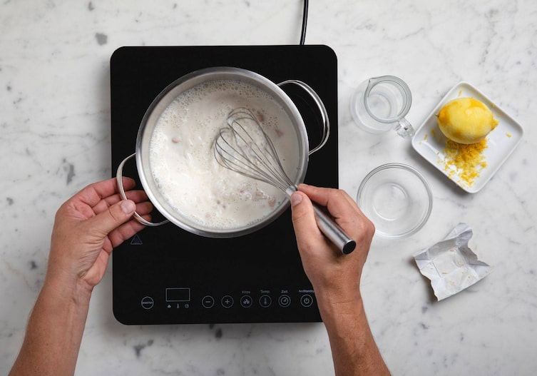 Kleine Kochplatte auf der ein Topf mit Milch steht und Germ eingerührt wird mit einem Schneebesen.