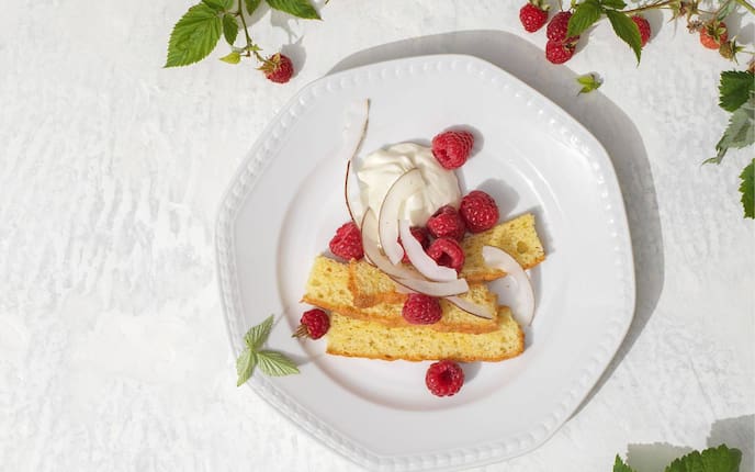 Feine Kuchenstreifen mit einer Creme und Himbeeren und Kokosstreifen auf einem weißen Keramikteller angerichtet