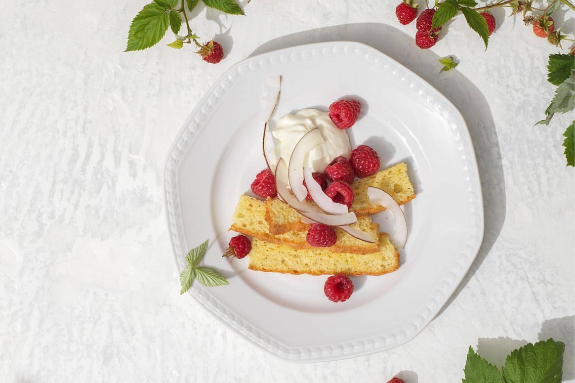 Feine Kuchenstreifen mit einer Creme und Himbeeren und Kokosstreifen auf einem weißen Keramikteller angerichtet