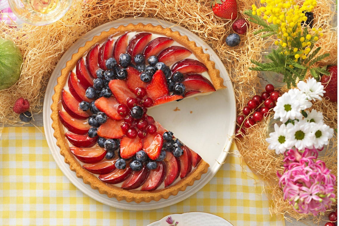 Auf einem Tisch mit gelb kariertem Tischtuch liegt Holzwolle, darauf ist frisches Beerenobst verstreut und Sommerblumen. Mitten drin steht ein weißer runder Teller mit einem flachen Kuchen, der mit Pfirsichen und verschiedenen Beeren aufgefächert gefüllt ist.