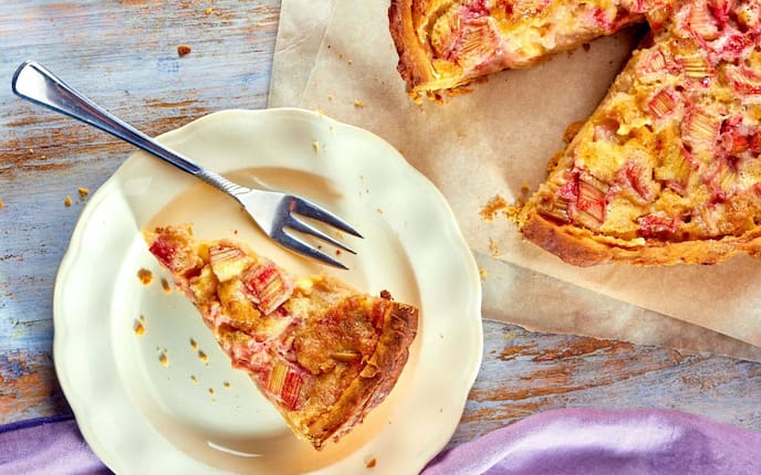 Rhabarberkuchen im Ganzen und ein Stück davon auf einem weißen Keramikdessertteller mit einer Dessertgabel
