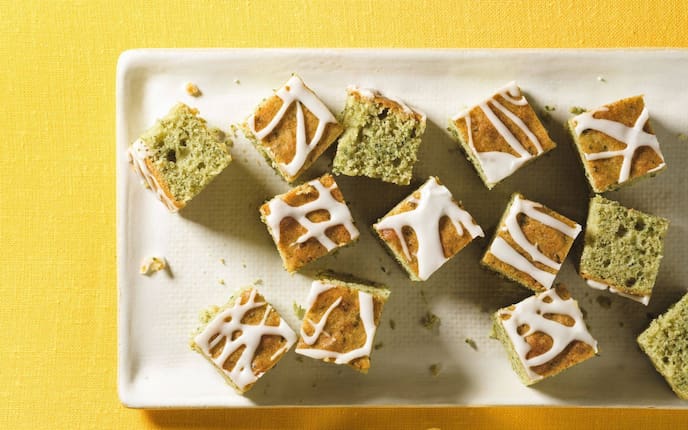 klein gewürfelter Blechkuchen, der etwas grünlich ist mit Zitronenguss auf einer weißen rechteckigen Platte auf einem quietschgelben Tischdecke