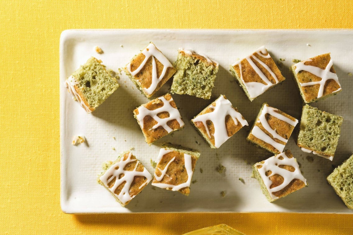 klein gewürfelter Blechkuchen, der etwas grünlich ist mit Zitronenguss auf einer weißen rechteckigen Platte auf einem quietschgelben Tischdecke
