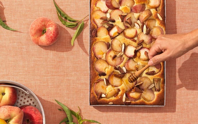 Ein Blech mit einem Pfirsich-Kuchen und Mandelstücken, eine Hand gießt mit einem Honiglöffel Honig darüber. Alles steht auf eine lachsfärbigen Tischtuch mit einem Alusieb, das mit frischen Pfirsichen gefüllt ist