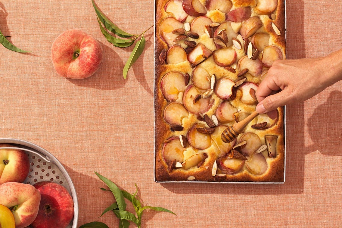 Ein Blech mit einem Pfirsich-Kuchen und Mandelstücken, eine Hand gießt mit einem Honiglöffel Honig darüber. Alles steht auf eine lachsfärbigen Tischtuch mit einem Alusieb, das mit frischen Pfirsichen gefüllt ist
