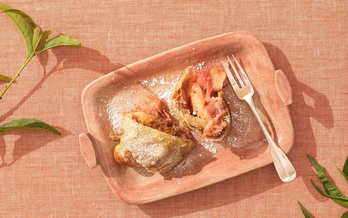 Ein lachsfärbiges Tischstuch auf dem ein lachsfärbiger eckiger Teller mit zwei Stück Pfirisichstrudel und einer Dessertgabel.