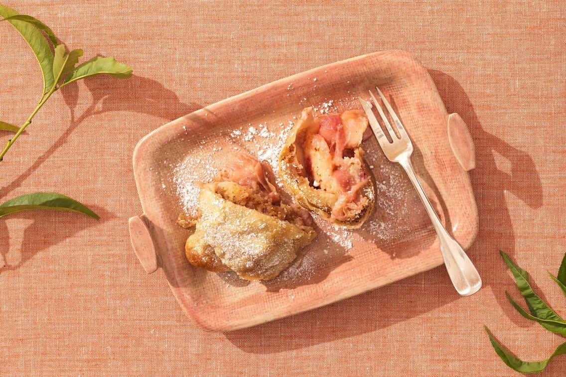 Ein lachsfärbiges Tischstuch auf dem ein lachsfärbiger eckiger Teller mit zwei Stück Pfirisichstrudel und einer Dessertgabel.