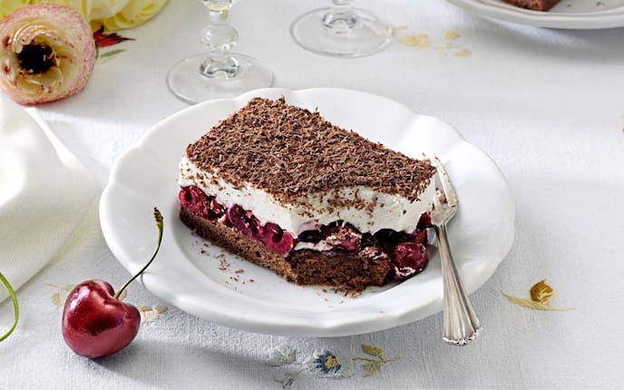 Ein Stück Cremeschnitte mit Schokoteig und Kirschen auf einem weißen Teller. Alles zusammen steht auf einem festlich gedeckten Tisch.