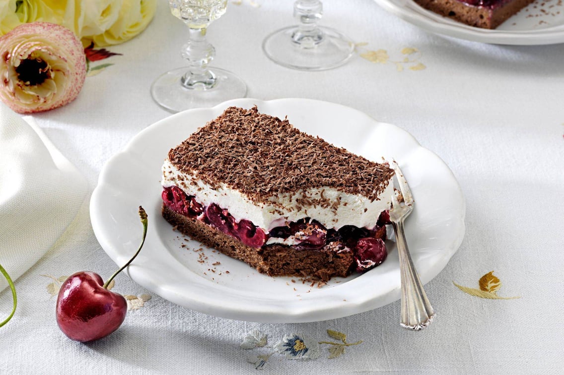 Ein Stück Cremeschnitte mit Schokoteig und Kirschen auf einem weißen Teller. Alles zusammen steht auf einem festlich gedeckten Tisch.