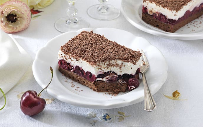 Ein Stück Cremeschnitte mit Schokoteig und Kirschen auf einem weißen Teller. Alles zusammen steht auf einem festlich gedeckten Tisch.