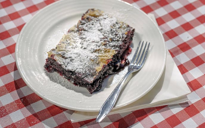 Ein Stück flacher Sturdel mit Schwarzbeeren gefüllt und mit Staubzucker bestreut, liegt auf einem weißen Teller neben einer Dessertgabel. Alles steht auf einem rot karierten Tischtuch und einer Serviette.