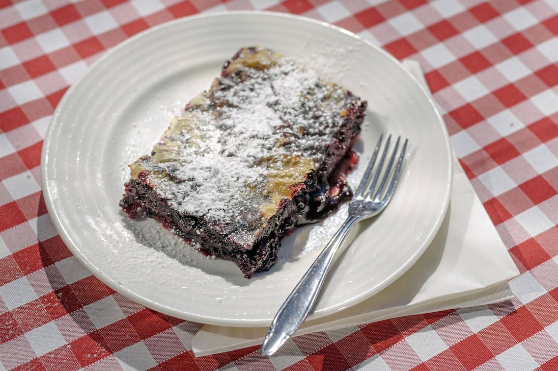 Ein Stück flacher Sturdel mit Schwarzbeeren gefüllt und mit Staubzucker bestreut, liegt auf einem weißen Teller neben einer Dessertgabel. Alles steht auf einem rot karierten Tischtuch und einer Serviette.