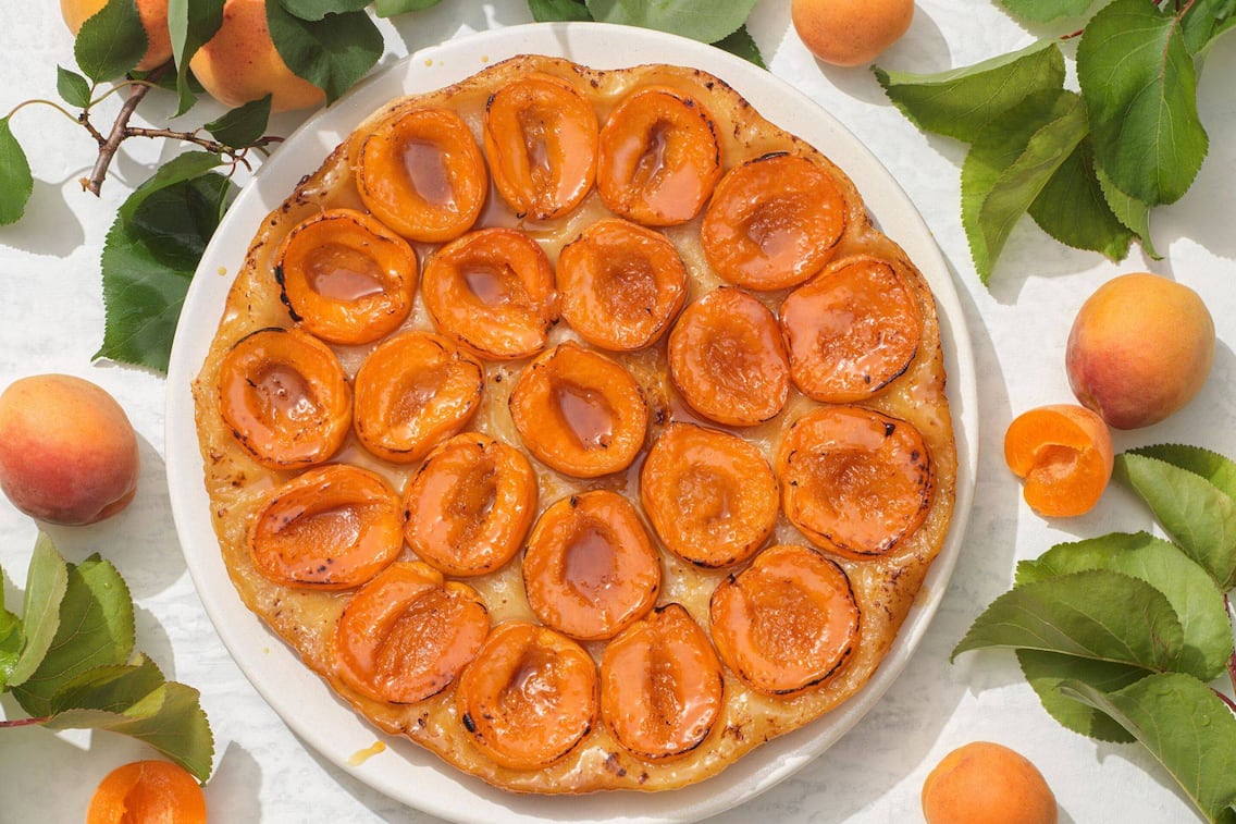 Eine Tarte mit Marillenhälften auf einem runden Keramikteller, darum herum liegen Blätter eines Marillenbaumes und frische Marillen.