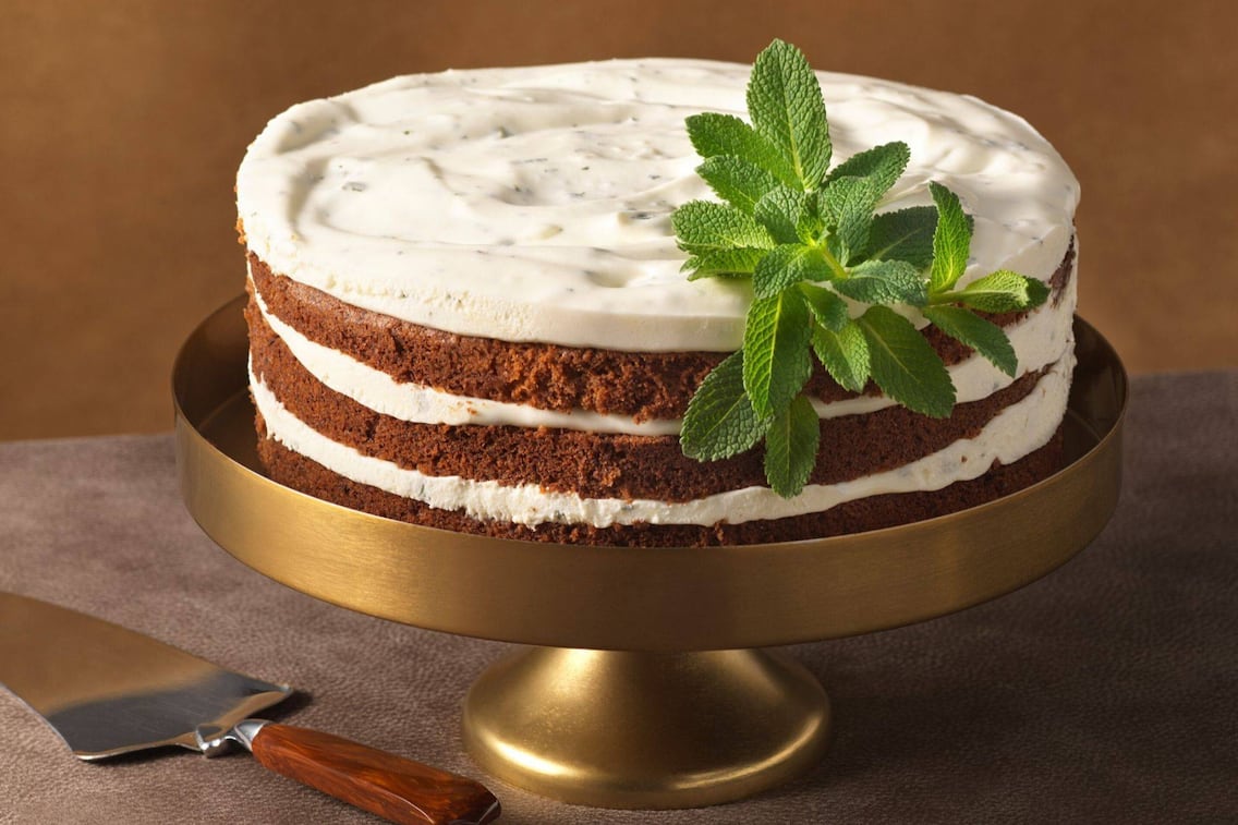 Eine geschichtete Topfencremetorte mit drei dunklen Teigböden und Topfenschichten. Obenauf ist auch eine Topfenschicht mit einem Minzezweig verziert. Die Torte steht auf einem niedrigen Tortenständer aus Messing, daneben liegt ein Tortenheber mit dunklem Holzgriff.