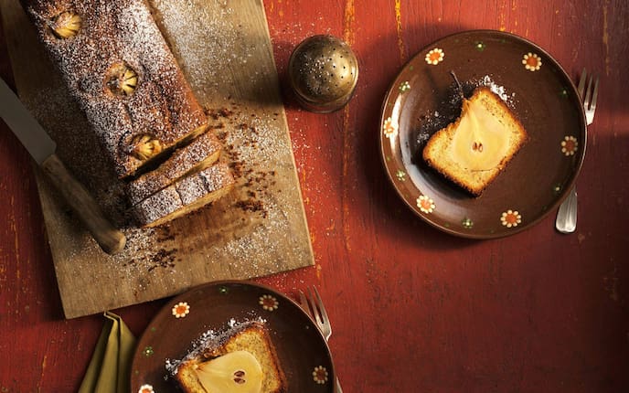 Heller Kastenkuchen mit einer ganzen Birne im Stück, Teile davon aufgeschnitten auf braunen Dessertellern angerichtet, man sieht die ganze Birne im Längsschnitt im Kuchen.