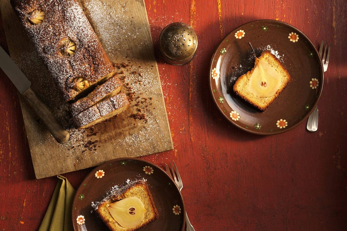 Heller Kastenkuchen mit einer ganzen Birne im Stück, Teile davon aufgeschnitten auf braunen Dessertellern angerichtet, man sieht die ganze Birne im Längsschnitt im Kuchen.