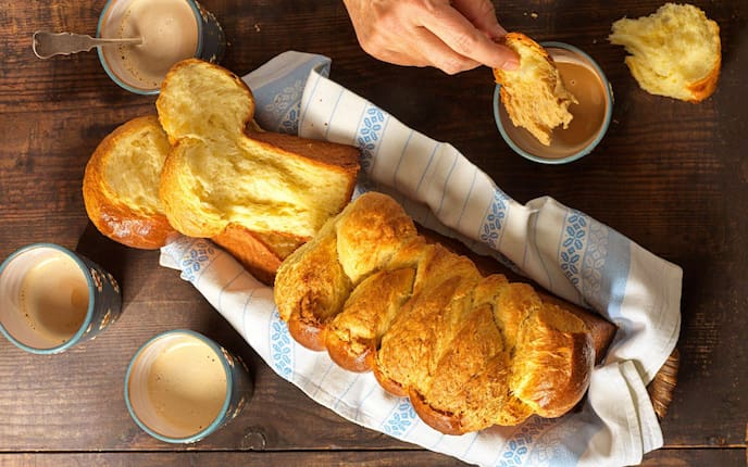 Ein ganzer Brioche in einem Weidekorb, in dem ein blau-weißes Geschirrtuch liegt. Daneben stehen vier Keramiktrinkbecher mit Kaffee und eine Hand tunkt ein Stück Brioche in den Kaffee.