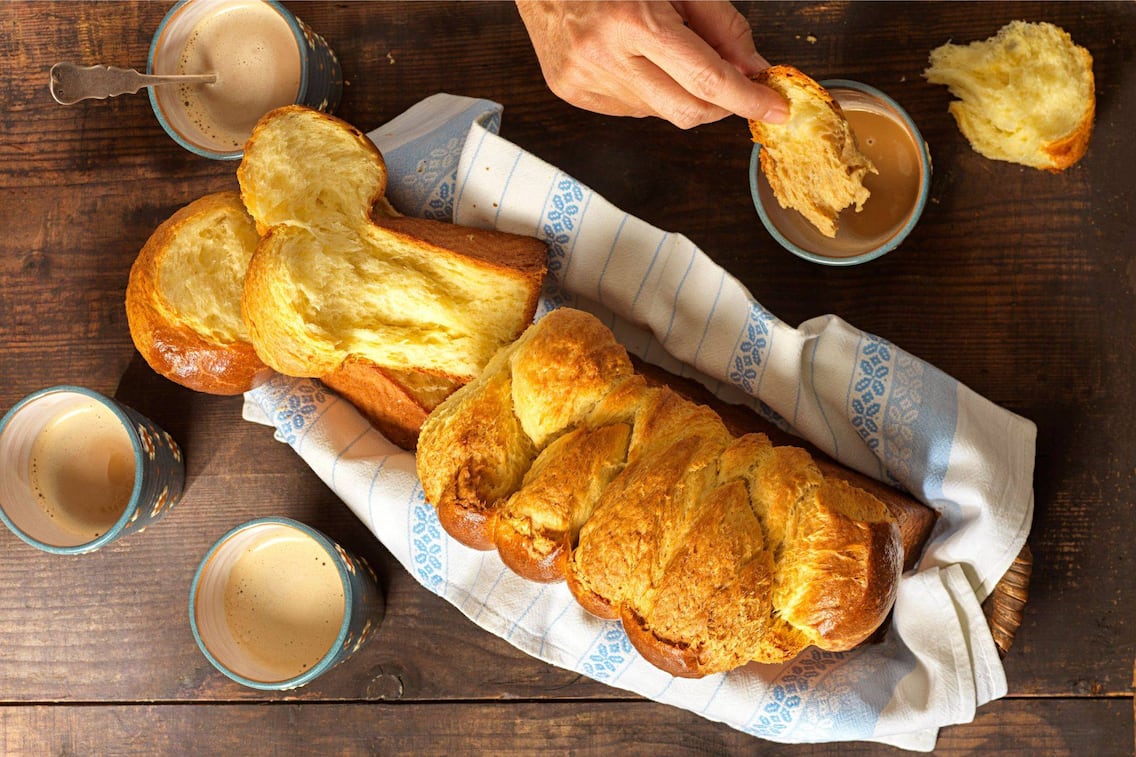 Ein ganzer Brioche in einem Weidekorb, in dem ein blau-weißes Geschirrtuch liegt. Daneben stehen vier Keramiktrinkbecher mit Kaffee und eine Hand tunkt ein Stück Brioche in den Kaffee.