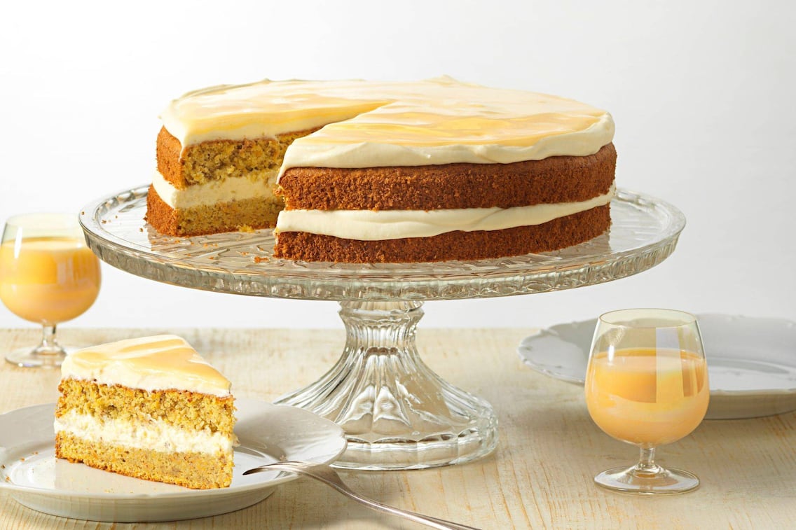 Torte mit Cremeschichte in der Mitte und Oben. Sie steht auf einem gläsernen Tortenhalter. Daneben steht ein Desserteller mit einem Stück der Torte und zwei gläser Eierlikör.