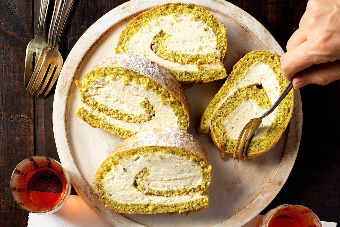 Grüne Biskuitroulade mit weißer Cremefüllung, in Scheiben geschnitten auf einem Holzteller. Man sieht eine männliche Hand, die mit einer Dessertgabel in ein Stück sticht. Alles steht auf einem dunkelbraunen Holzuntergrund. Darauf stehen zwei kleine Weingläser mit Schilcher gefüllt und drei Dessertgabeln liegen auch noch da.