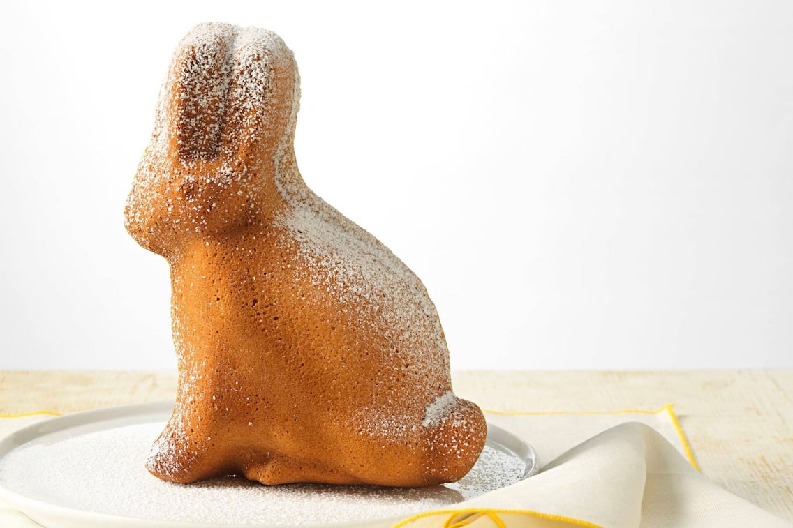 Mit Staubzucker bestreuter Osterhase in Kuchenform auf einem weißen Teller, der auf einem weißen Tischtuch mit gelben Rand steht auf einem weißen Holztisch.