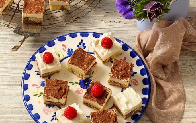 Topfen-Biskuitküchlein in Würfel geschnitten und mit Himbeeren dekoriert, leigen auf einem blau-weißen Blumenteller, der auf einem Holztisch steht, daneben liegt eine braune Stoffserviette.