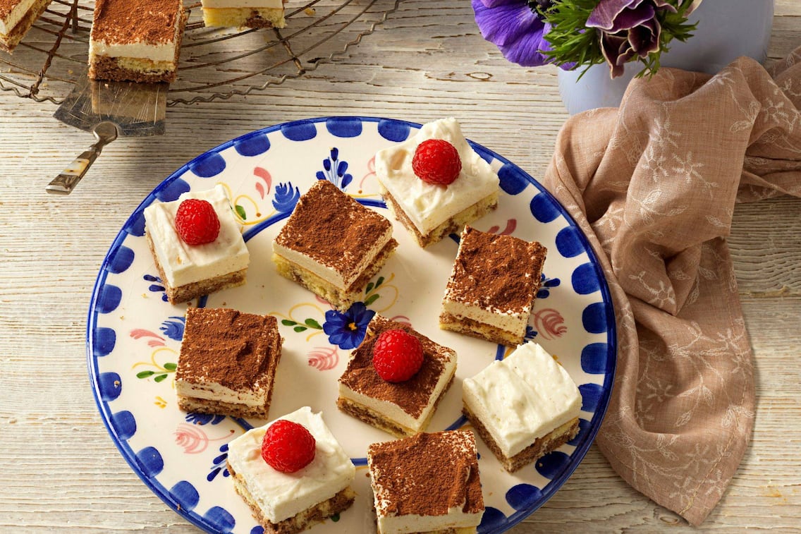 Topfen-Biskuitküchlein in Würfel geschnitten und mit Himbeeren dekoriert, leigen auf einem blau-weißen Blumenteller, der auf einem Holztisch steht, daneben liegt eine braune Stoffserviette.