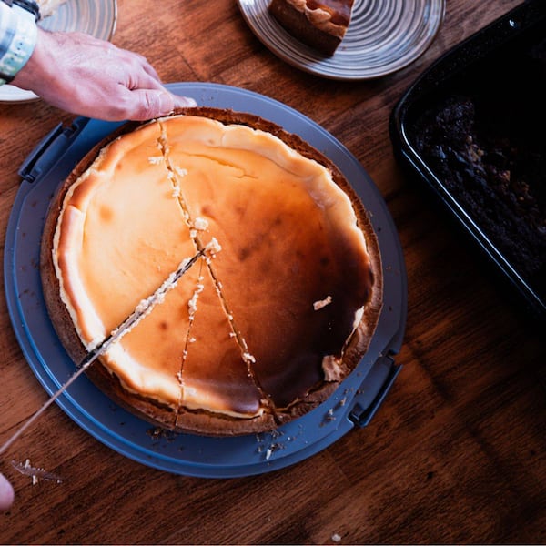 Topfentorte steht auf dem Unterteil einer blauen Tortentransportform, Männerhände schneiden mit einem großen Messer Torgenstücke von der Torte. Alles steht auf einem dunkelbraunen Holztisch