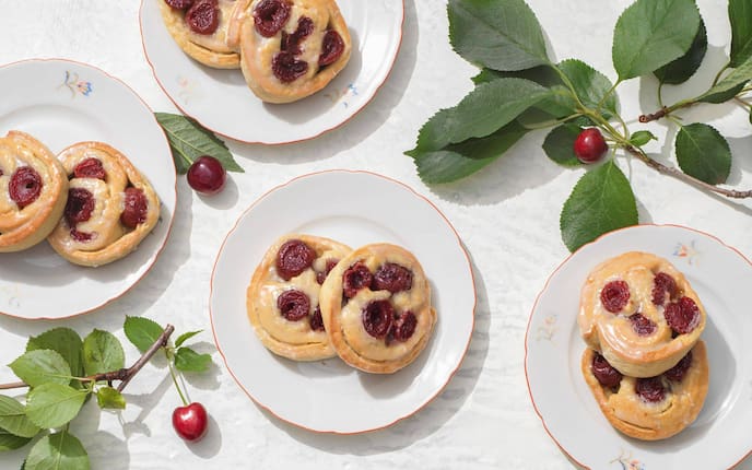 auf kleinen Desserttellern liegen jeweil zwei Germteigschnecken mit Kirschhälften. Daneben liegen frische Kirschzweige mit Kirschen.