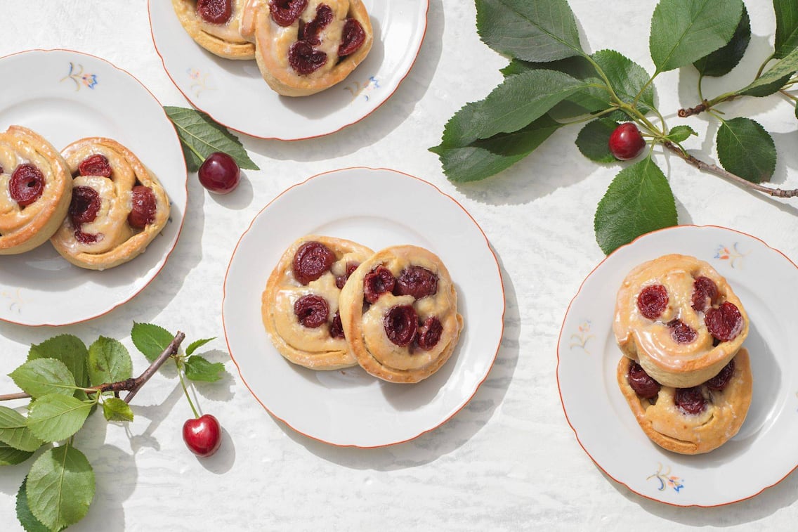 auf kleinen Desserttellern liegen jeweil zwei Germteigschnecken mit Kirschhälften. Daneben liegen frische Kirschzweige mit Kirschen.