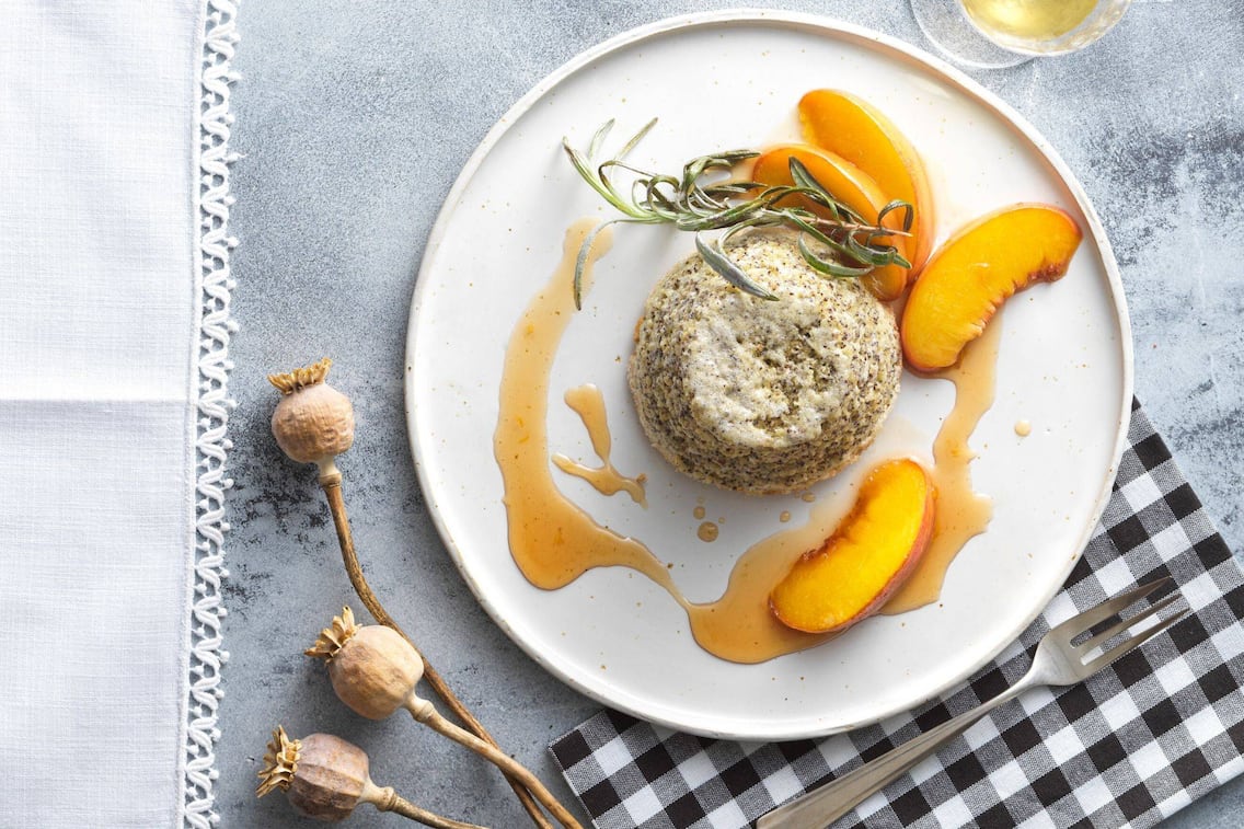 Mohnauflauf mit Ahornsirup und Pfirsichspalten und einem Lavendelzweig auf einem weißen Teller angerichtet.